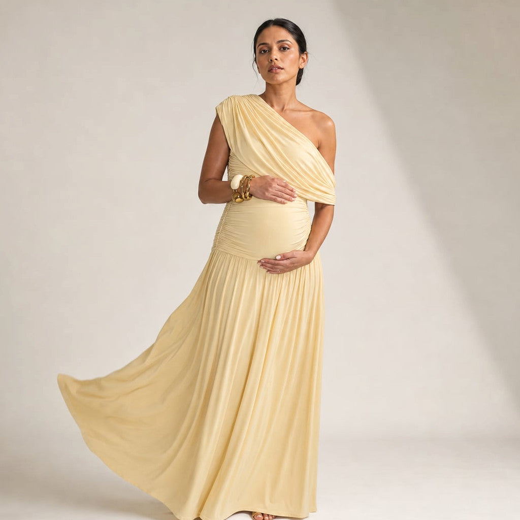 Model wearing Rowan asymmetric one-shoulder maternity maxi dress in apricot, showing bump-skimming silhouette and diagonal neckline